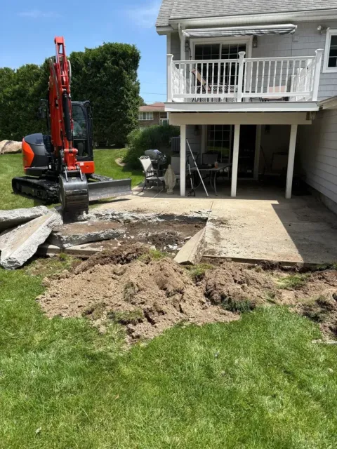 Excavator digging up concrete in front of a white house with a balcony and open garage