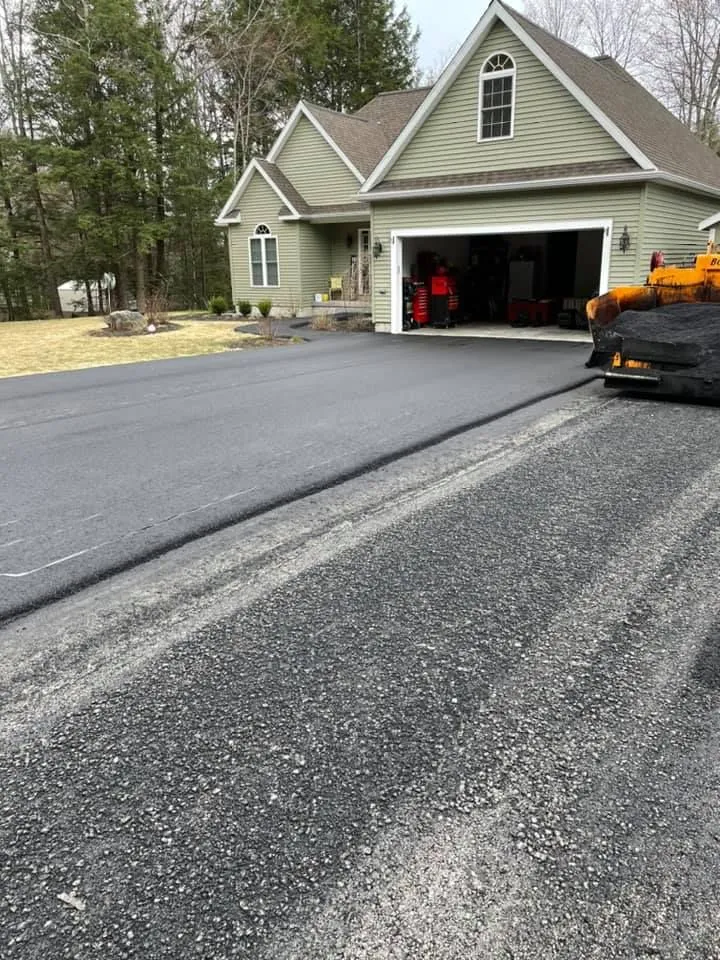 A light green house with a garage and a newly paved asphalt driveway in front of a gravel area.