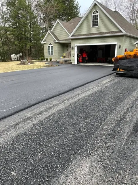 A light green house with a garage and a newly paved asphalt driveway in front of a gravel area.