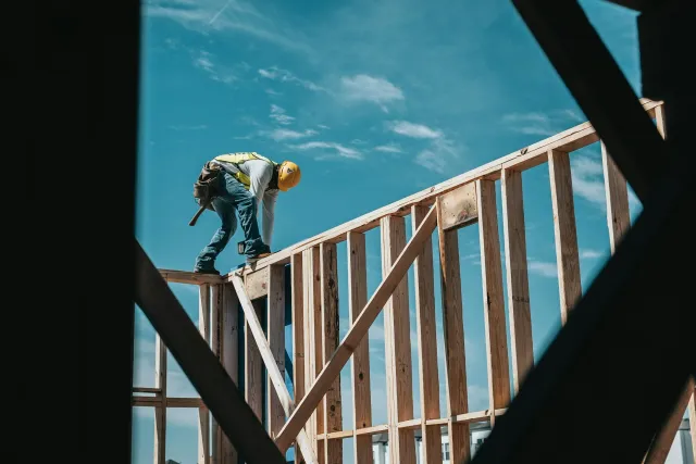A construction worker wearing a yellow hard hat and safety vest is working on the wooden framework of a building under a blue sky.
