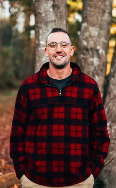 Man wearing red and black plaid shirt, smiling outdoors, with trees in the background.