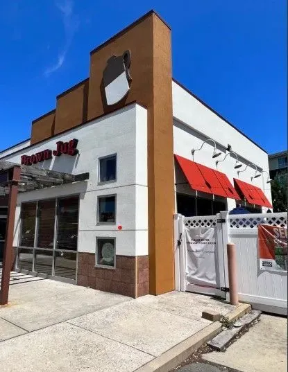 Exterior of a two-story restaurant building; white, brown, and orange facade; red awnings; sunny day.