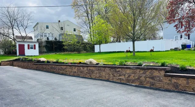 A stone retaining wall bordering a paved driveway, with a grassy lawn, trees, a shed, and houses in the background.