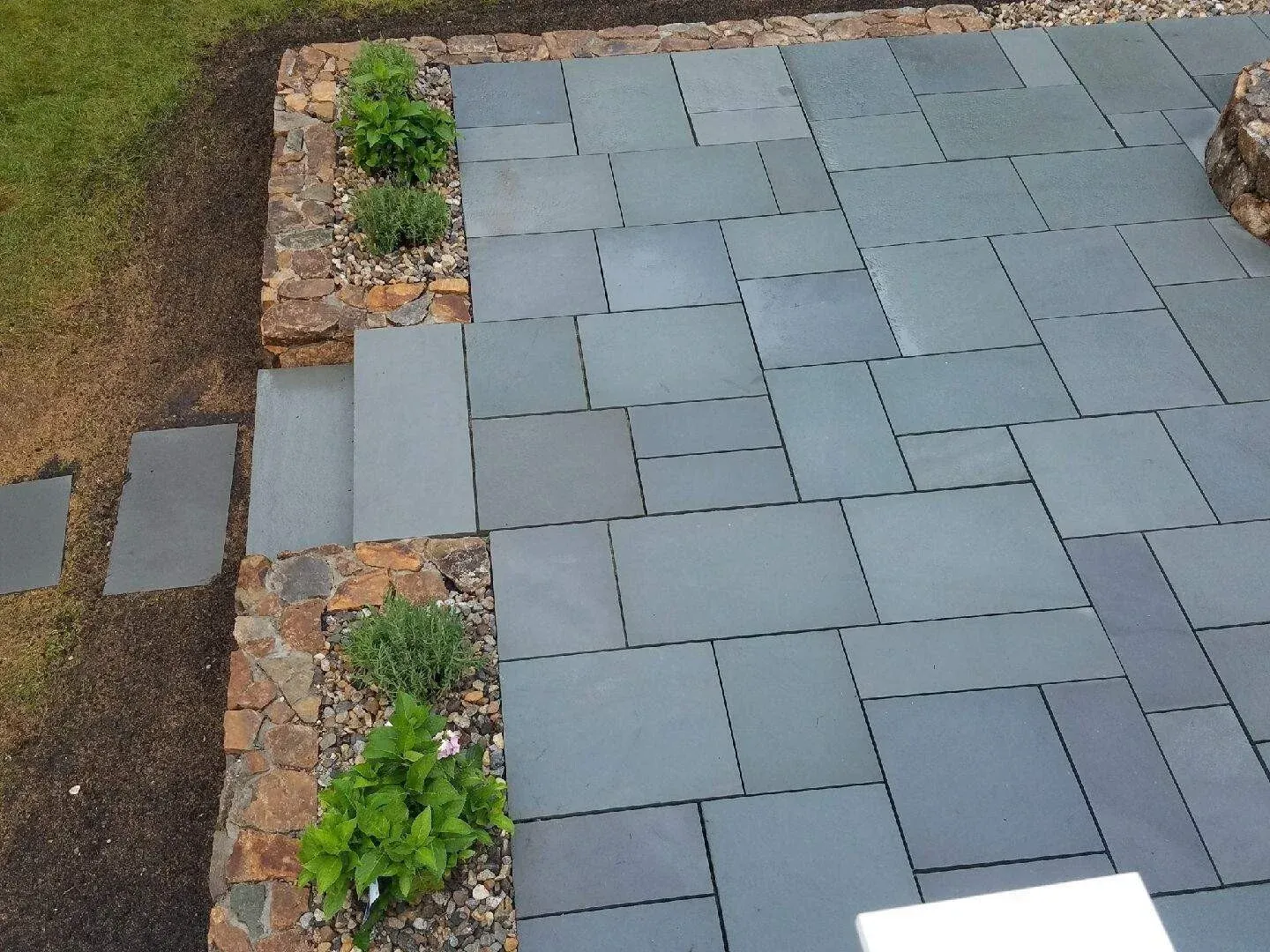 Newly laid gray stone pavers with a small flower bed and plants on the side, bordered by stone edging, and a patch of soil and grass beside it.