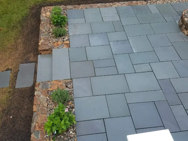 Newly laid gray stone pavers with a small flower bed and plants on the side, bordered by stone edging, and a patch of soil and grass beside it.