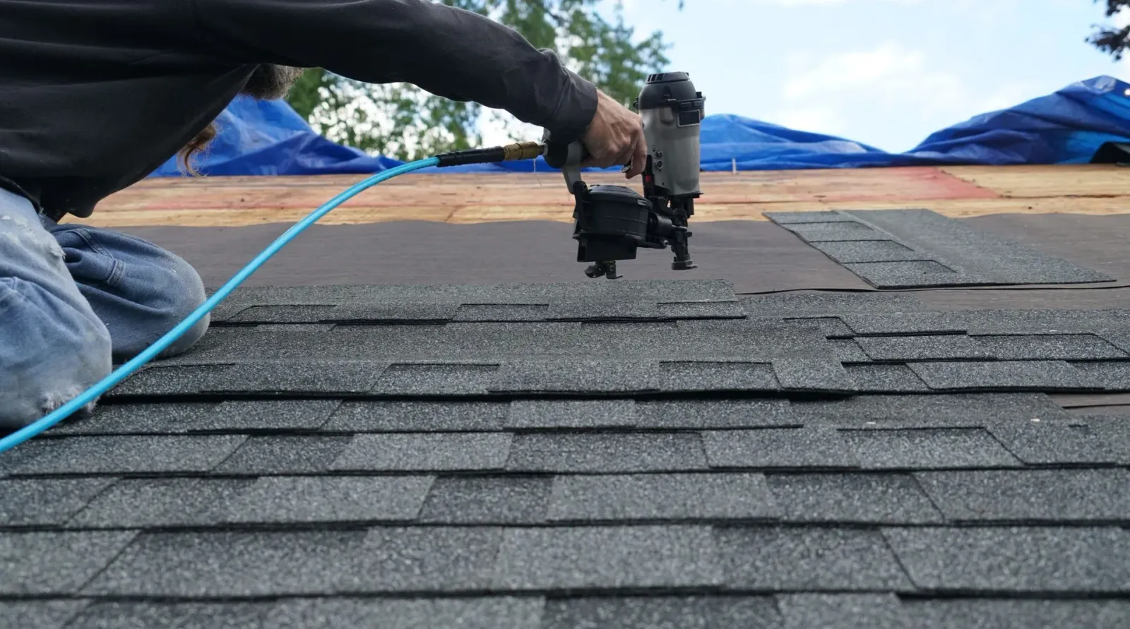 Roofer kneeling, using a nail gun to install asphalt shingles on a rooftop; blue air hose.