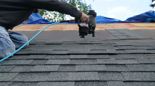 Roofer kneeling, using a nail gun to install asphalt shingles on a rooftop; blue air hose.