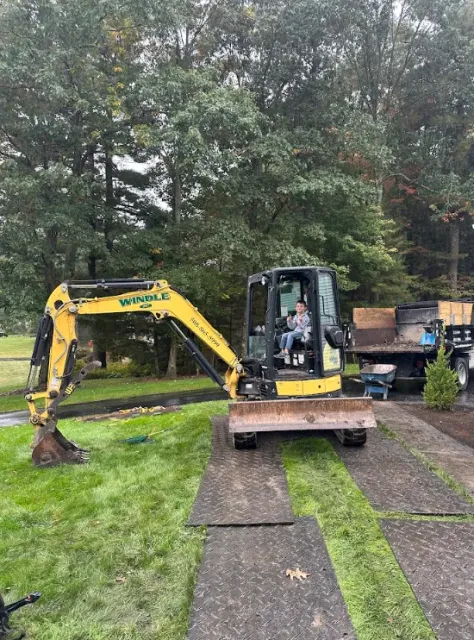 Yellow mini excavator on a lawn beside a driveway, with trees in the background.