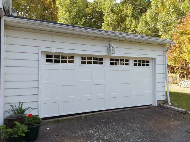 White detached garage with three doors and small windows, beside a driveway and autumn trees.