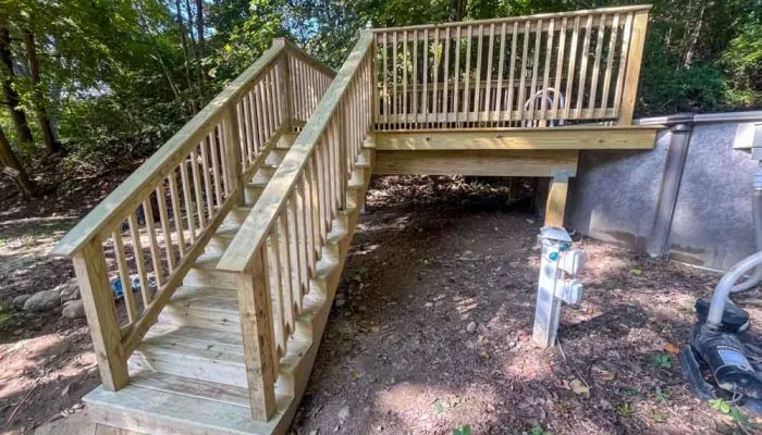 Outdoor wooden staircase with railings leading up to a small elevated deck platform beside a concrete foundation wall; surrounded by forest floor and greenery, showcasing rustic construction in progress.