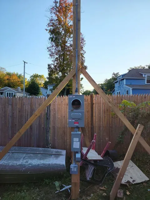Wooden utility pole with electric meter, braced by supports, beside a fenced backyard and wheelbarrow.