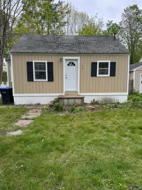 Small beige house with black shutters, white door, and overgrown yard.