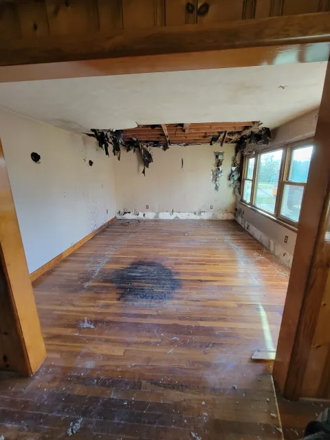 Damaged room with a large hole in the ceiling, soot on the walls, and burn marks on the floor, with peeling wallpaper and broken ceiling drywall.