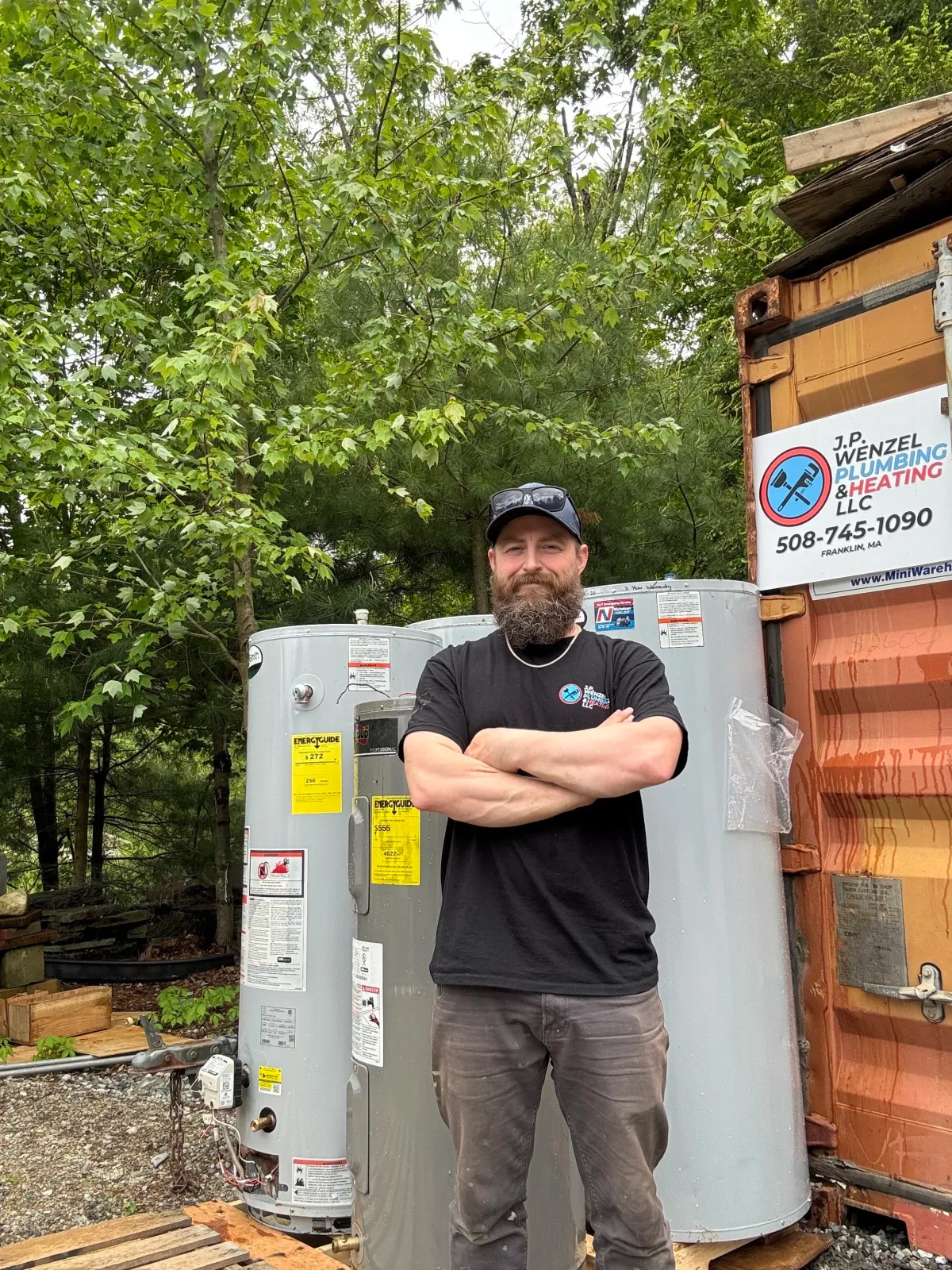 A man with a beard and sunglasses on his cap, dressed in a black T-shirt and gray pants, stands outdoors with his arms crossed in front of two water heaters and a wooden shed. Trees surround the area.