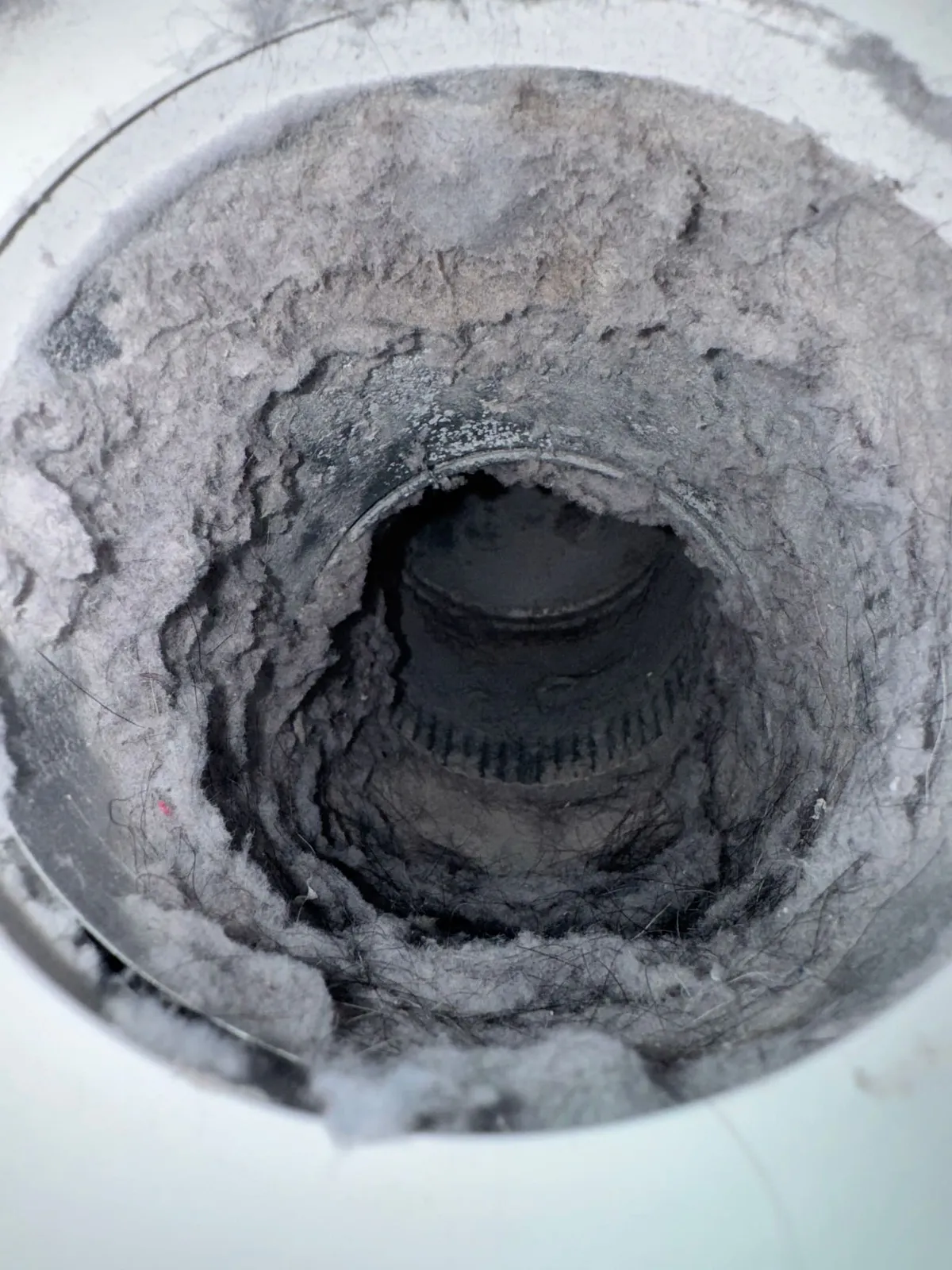 View down a dryer vent duct where compacted grey lint has narrowed the passage to a small dark opening.