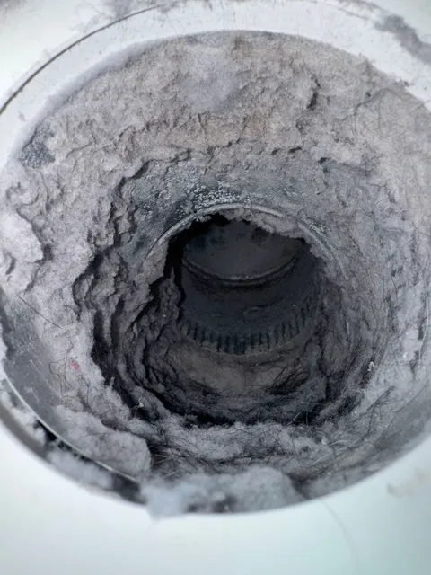 View down a dryer vent duct where compacted grey lint has narrowed the passage to a small dark opening.