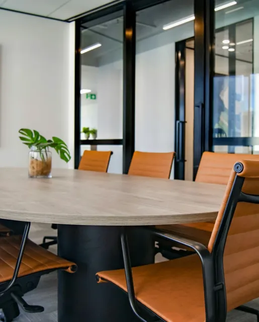 Modern conference room with wooden table, orange leather chairs, and a glass wall with black frames. A small potted plant is on the table.