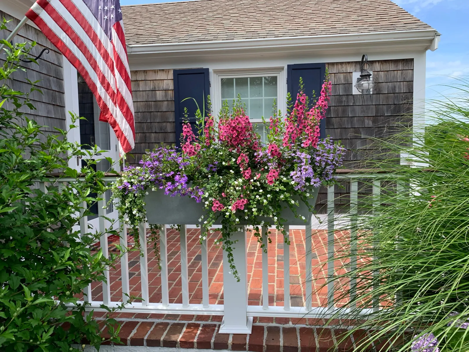 A house porch with a white railing and brick flooring, a window with navy blue shutters, a hanging lantern, pink and purple flowers in a white planter, and an American flag hanging on the left.