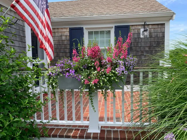 A house porch with a white railing and brick flooring, a window with navy blue shutters, a hanging lantern, pink and purple flowers in a white planter, and an American flag hanging on the left.