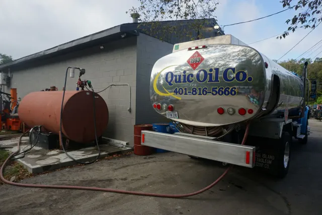 A Quic Oil Co. tanker truck parked by a building, pumping fuel into a large, cylindrical, reddish-brown storage tank.
