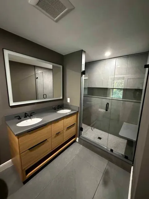 Modern bathroom with gray tile, wood vanity, glass shower, and rectangular mirror.