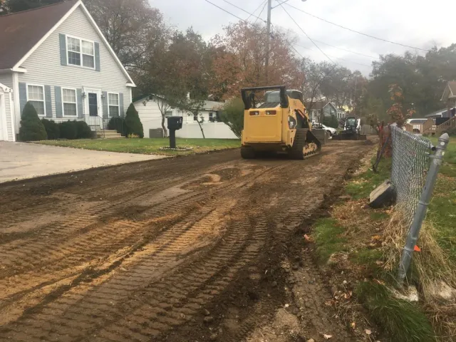 A skid steer tractor on a muddy road; residential houses in the background.
