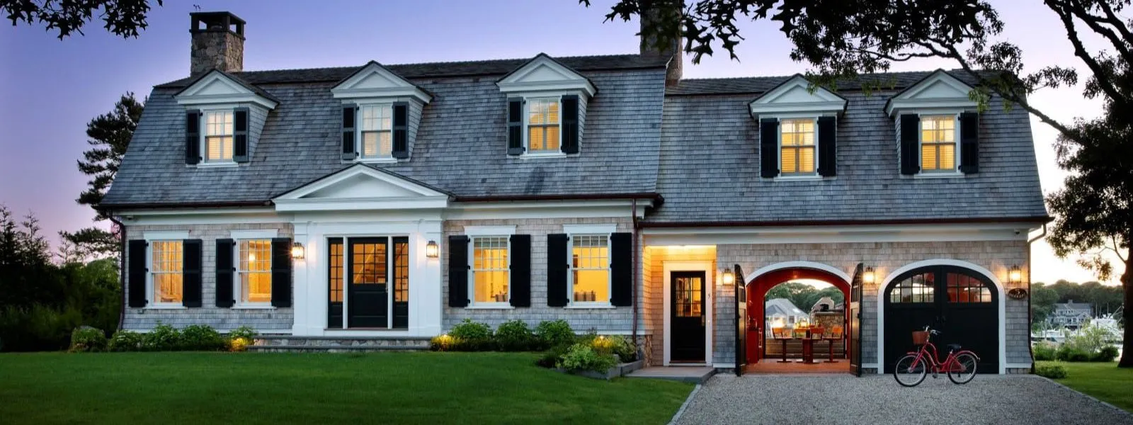 Two-story gray house with front porch, garage doors open, and red bike outside – McPhee Associates of Cape Cod.