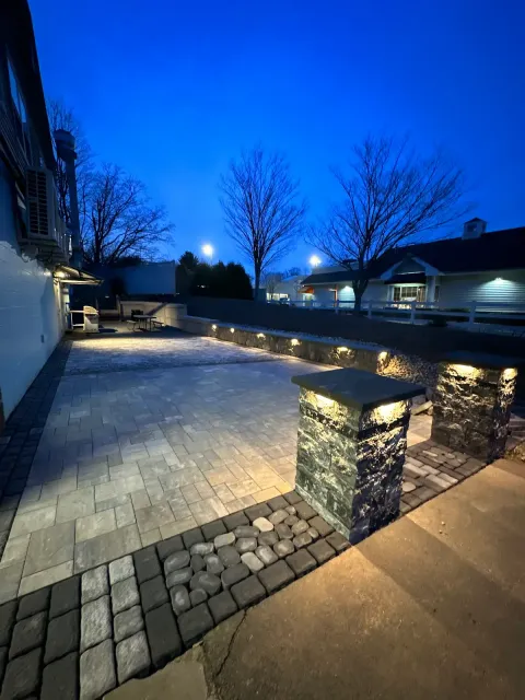 A paved backyard patio area at dusk with outdoor lighting, bordered by a stone wall, trees without leaves, and neighboring houses illuminated.