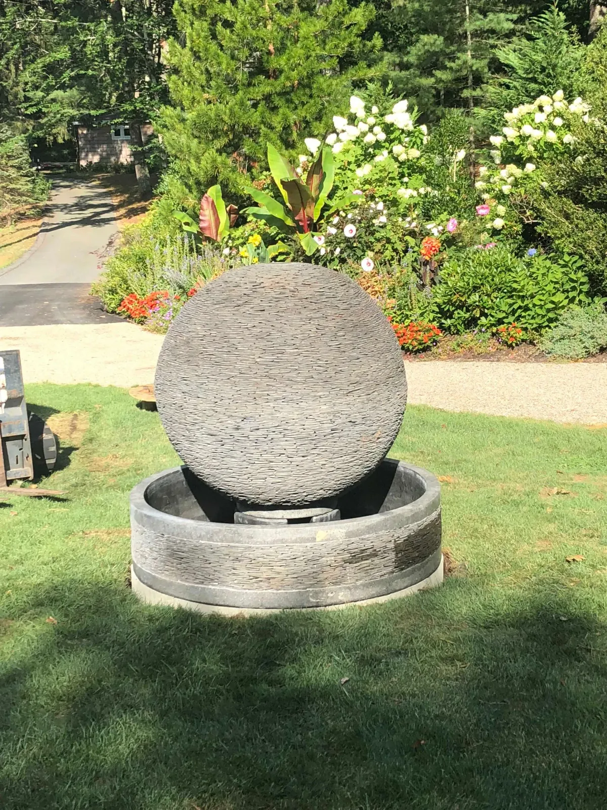 A large, textured stone sphere water feature sits in a green garden bed, centered in front of lush bushes and a path.