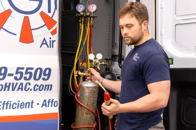'n Man in 'n blou hemp bedien HVAC-toerusting terwyl hy 'n gassilinder en meters naby 'n dienswa met kontakinligting en handelsmerk sigbaar hou.