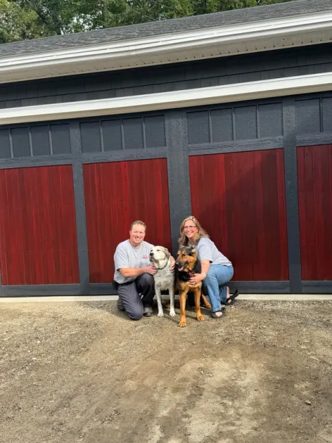 Two people kneel with two dogs in front of a barn with red doors.