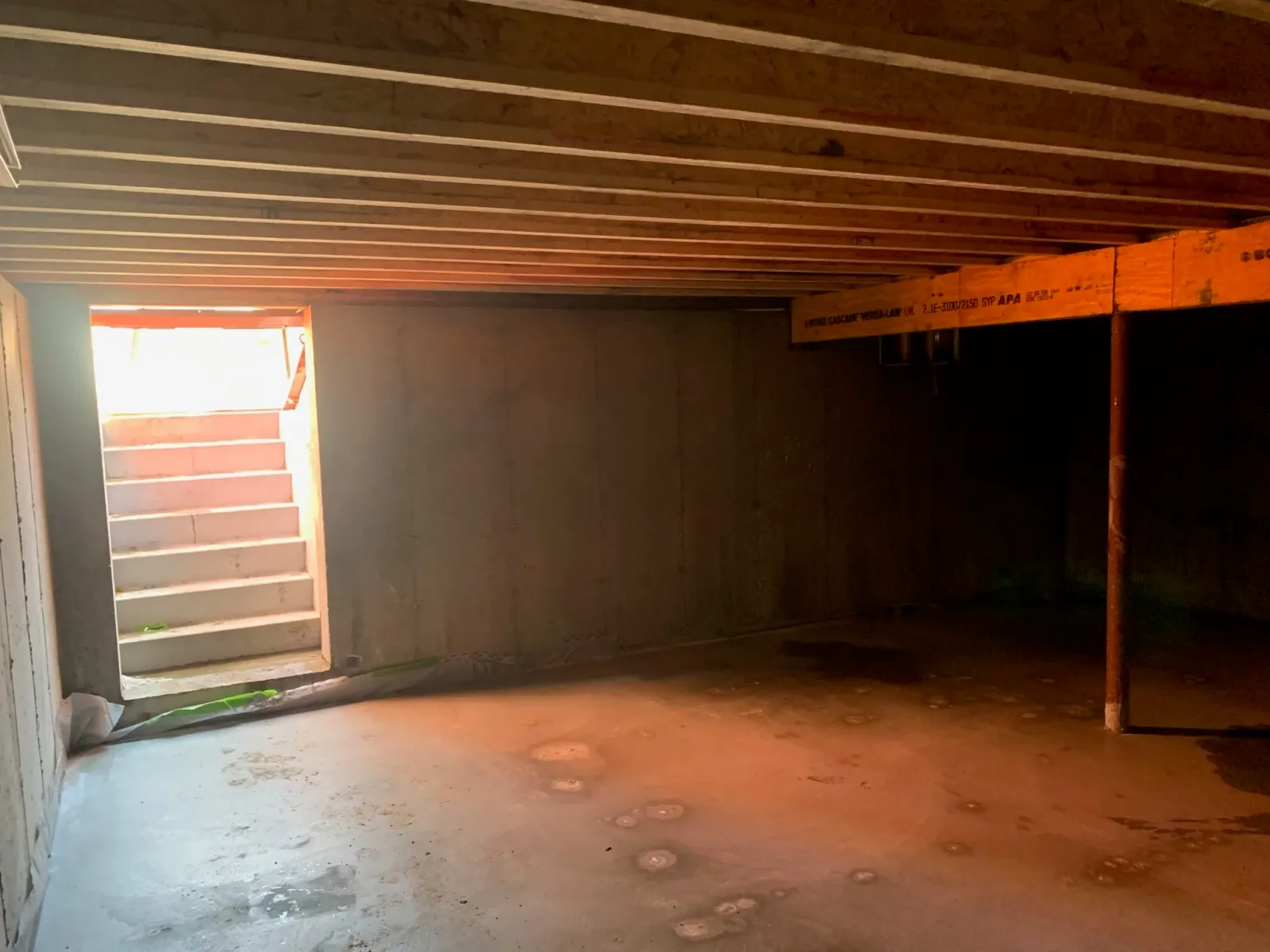 Bare interior of a building under construction with wooden ceiling beams, concrete walls, and a set of stairs leading outside, with light coming from the open doorway. Scituate, Norwell, Hanover, Hanson, Marshfield, South Shore. Massachusetts Local.
