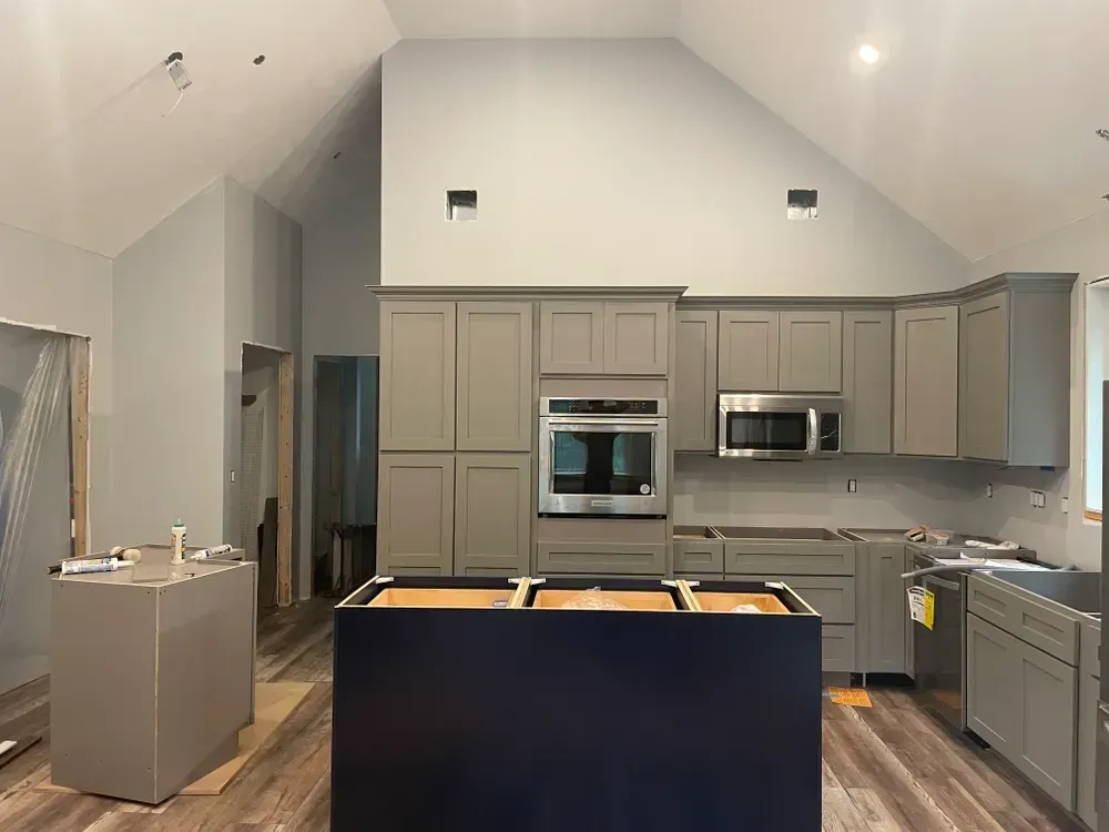 A kitchen under construction with gray cabinets, a blue island base, wood-look flooring, and a high, sloped ceiling.