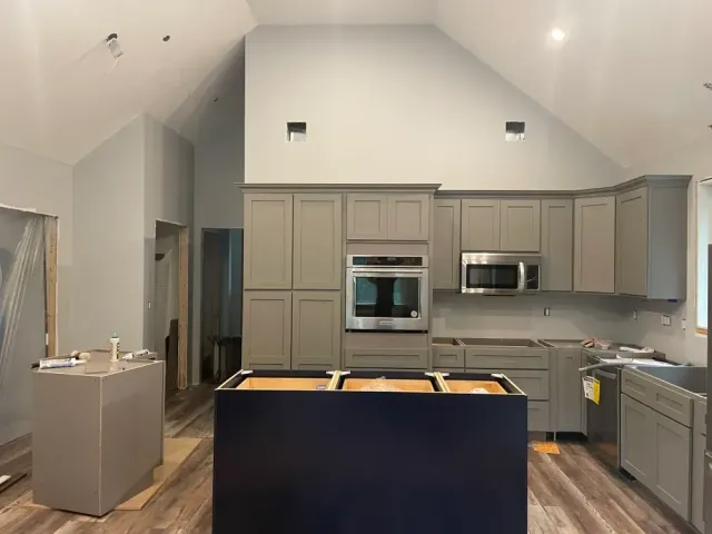 A kitchen under construction with gray cabinets, a blue island base, wood-look flooring, and a high, sloped ceiling.