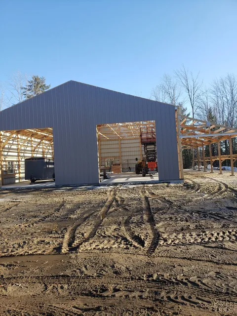 A steel pole barn under construction in Denmark, Maine, with the frame and trusses exposed through the open bay and a scissor lift inside
