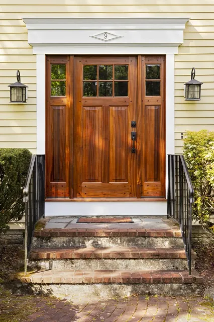 Front door of a house with a wooden door, two black lantern-style lights, a white decorative trim with the number 9, and steps leading up from a brick-paved porch.