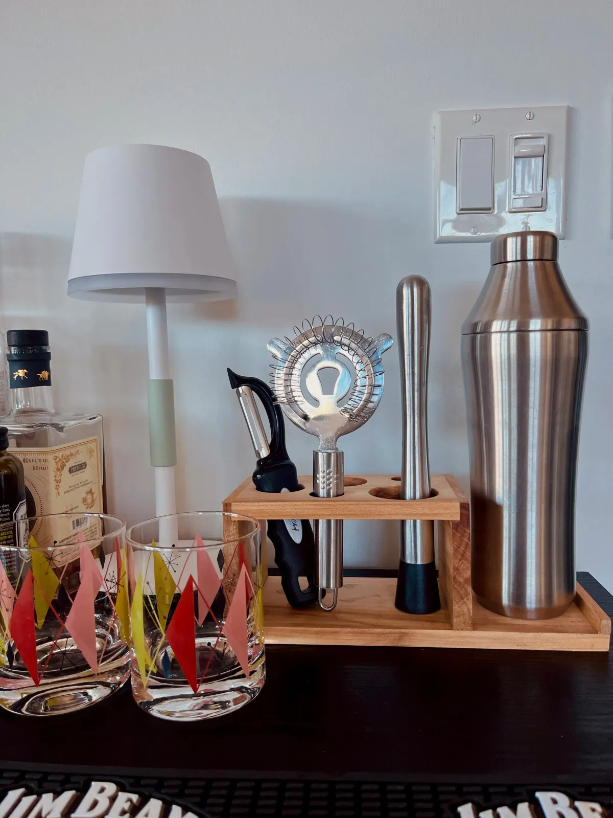Bar cart with a small white lamp, cocktail shaker, bar tools in a wooden holder, glasses, and bottles against a white wall.