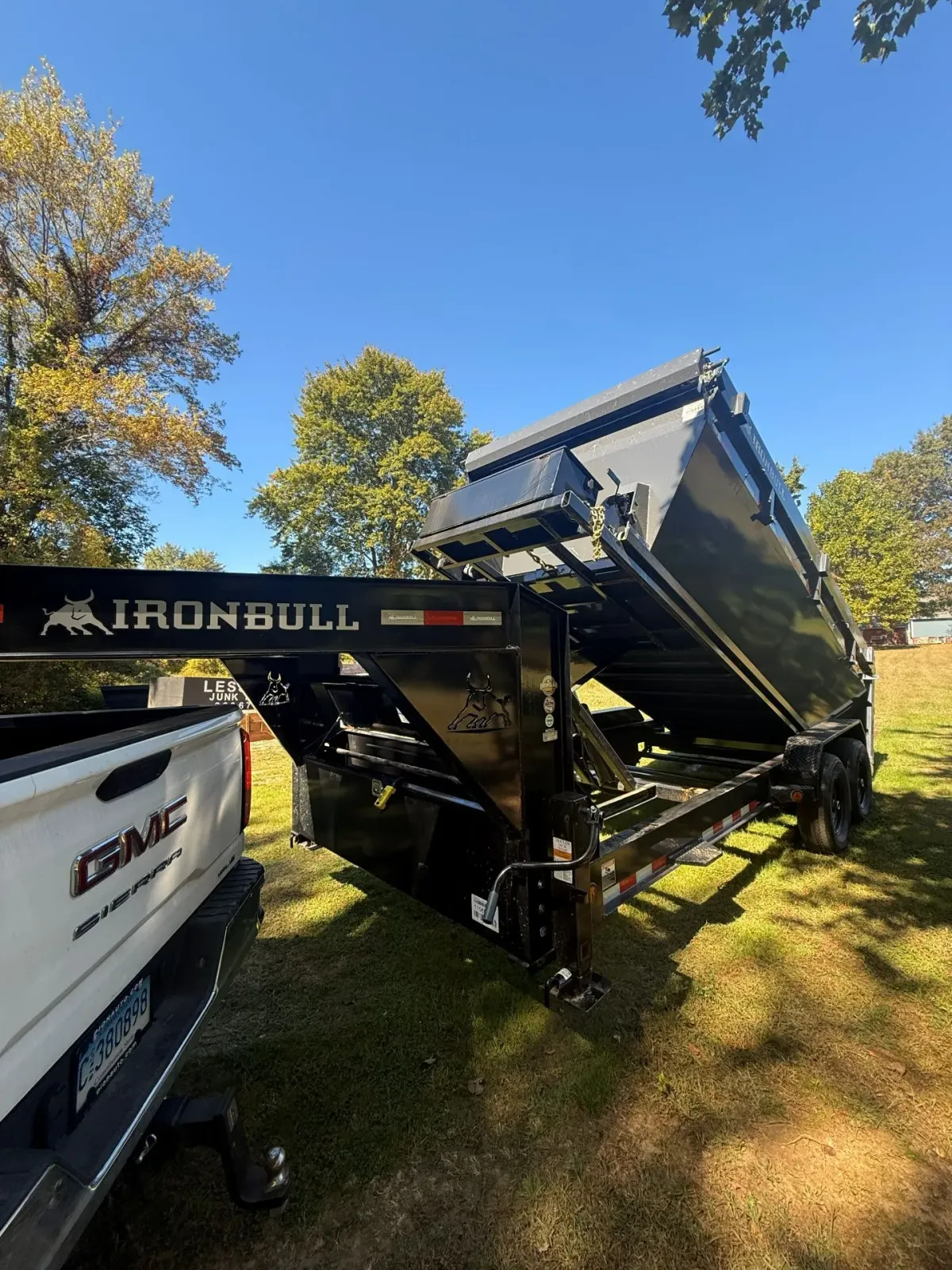 Black Ironbull dump trailer attached to a white GMC truck, raised bed, parked on grass, blue sky.