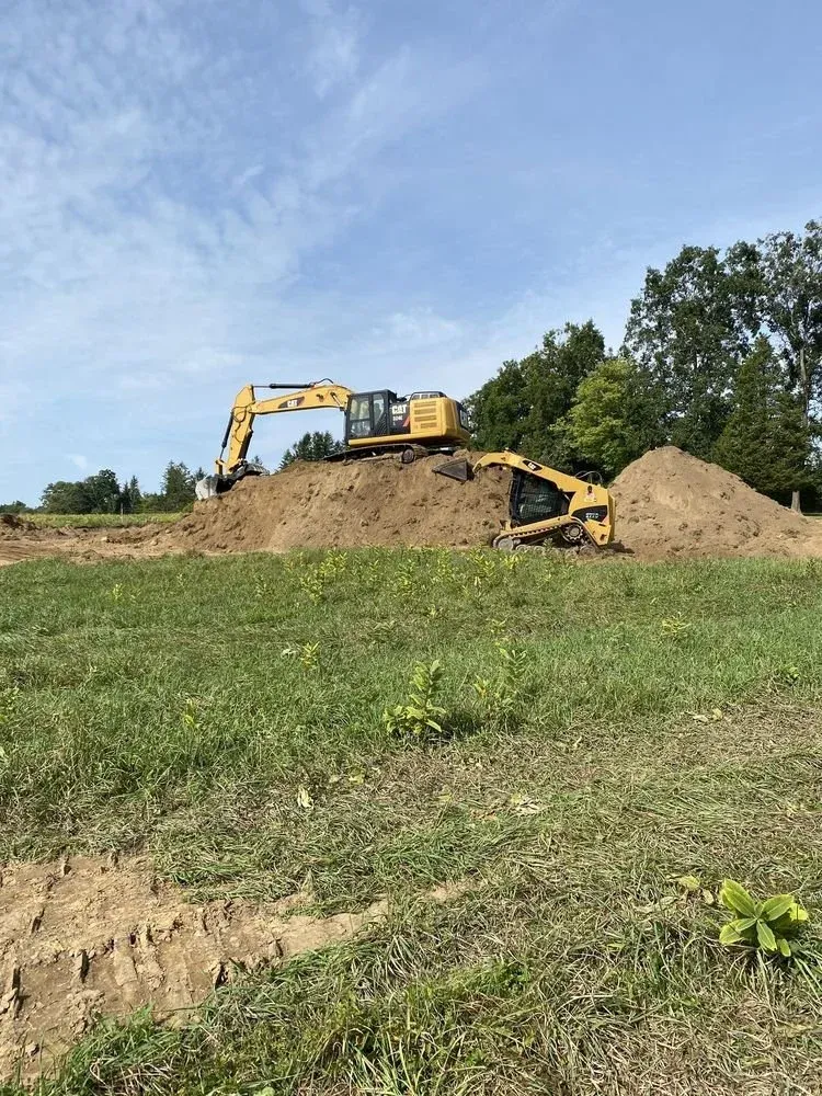 Construction site with two yellow excavators working on large mounds of dirt, surrounded by green grass and trees, under a partly cloudy sky.