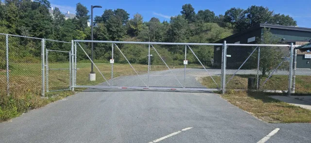 Closed metal gate blocking entrance to a parking lot or path, with trees and hillside in the background.