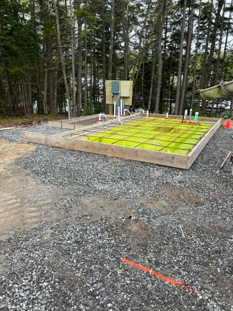 Construction site with wooden frame, rebar, and green plastic in-ground preparation, surrounded by gravel and dirt, with forest in background.