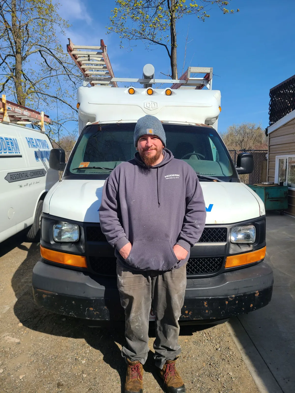 Person in a gray hoodie standing in front of a white utility van on a sunny street.