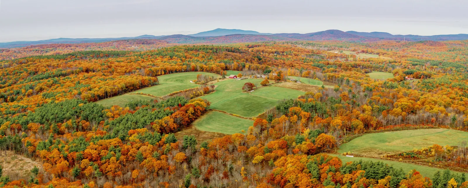 Aerial view of a red barn amid green fields surrounded by dense autumn forest with distant mountains