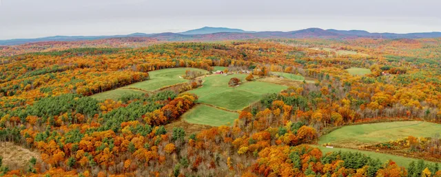 Aerial view of a red barn amid green fields surrounded by dense autumn forest with distant mountains
