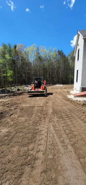 Construction site with a bulldozer grading dirt near a house, surrounded by trees and blue sky.