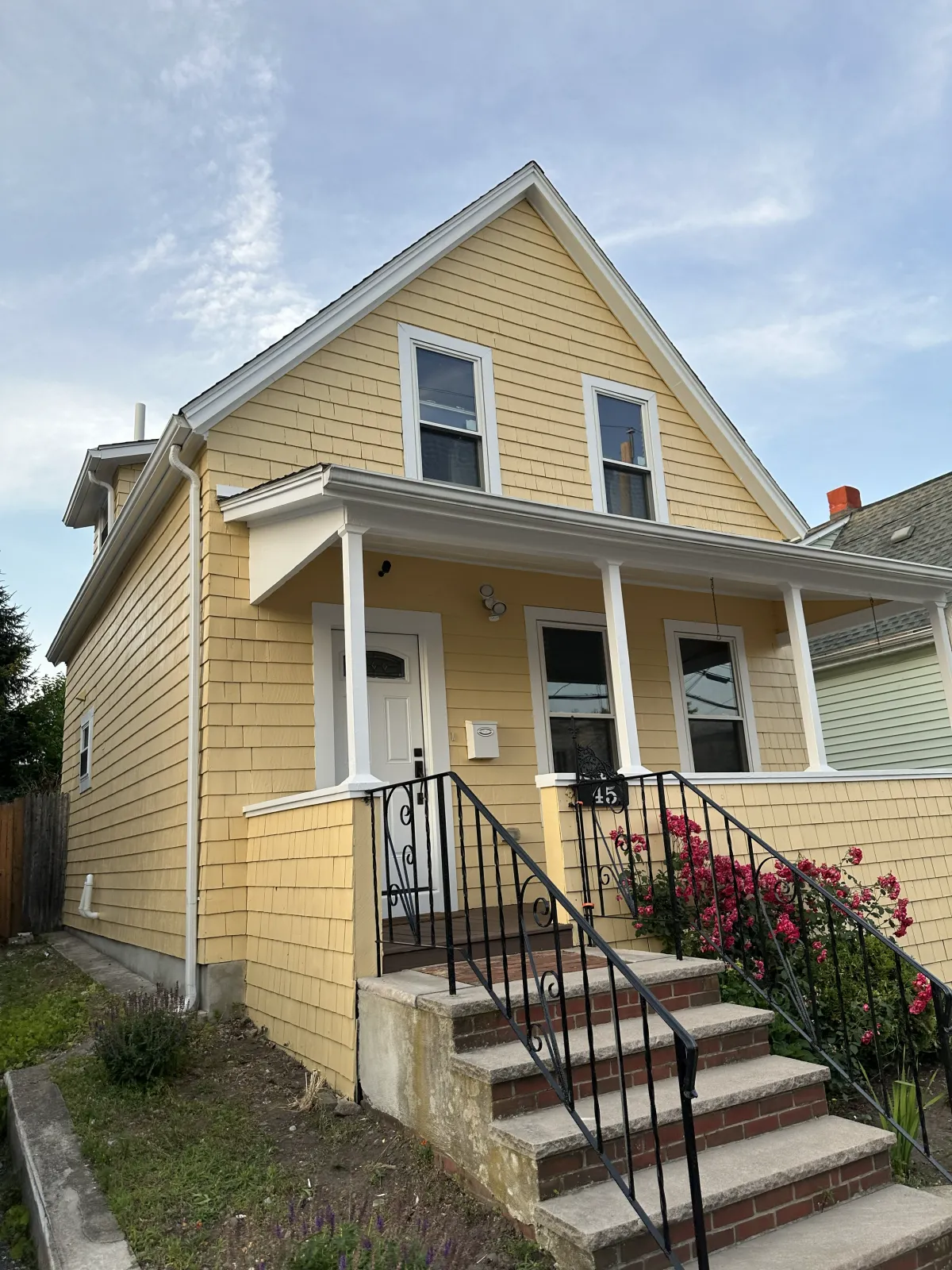 Yellow two-story house with a front porch, black railing, pink flowering bushes, and a pathway with stairs leading to the front door.
