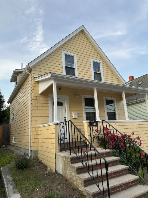 Yellow two-story house with a front porch, black railing, pink flowering bushes, and a pathway with stairs leading to the front door.