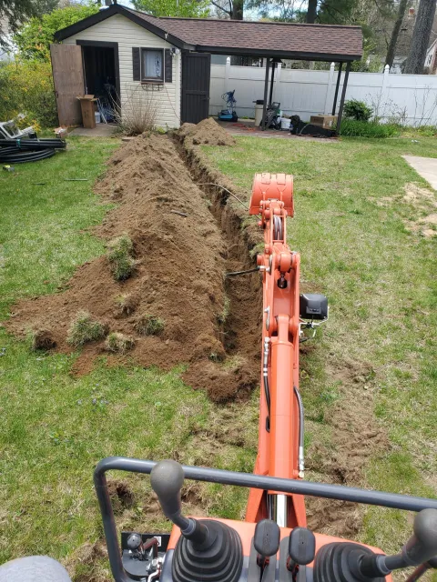 View from a construction vehicle, showing a freshly dug trench in a backyard, with a small shed and various tools and equipment around.