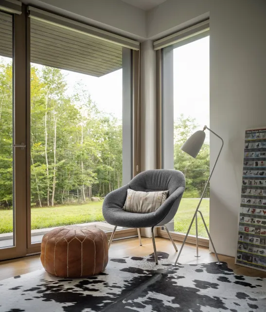 Modern living room with a large floor-to-ceiling window showing a green outdoor scene, a gray armchair with a decorative pillow, a hanging floor lamp, a leather pouf, a black-and-white cowhide rug, and a display rack with various items.