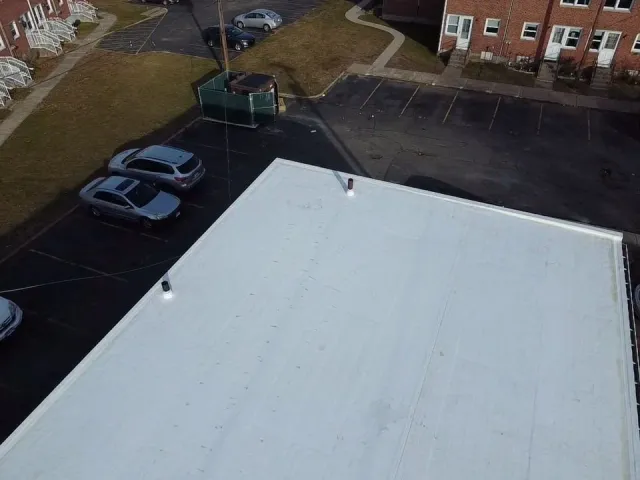 Aerial view of a white flat roof with two vents, parking lot with parked cars, residential buildings, a grassy area, walkways, and a chain-link fence.
